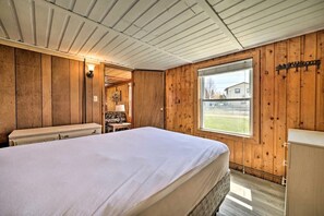 Une pièce comprenant un lit, une petite table, une lampe et une fenêtre donnant sur une maison.