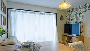 A dining area with a wooden table, chairs, a television, and a window with curtains. - La Réglisse - À moins d’1 km de la plage (Courseulles-sur-Mer)
