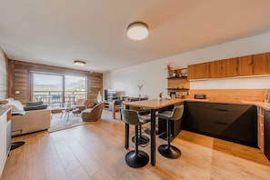 Eine moderne Küche mit Holzausstattung, ein Essbereich mit Barhocker und ein großes Fenster mit Blick ins Freie.