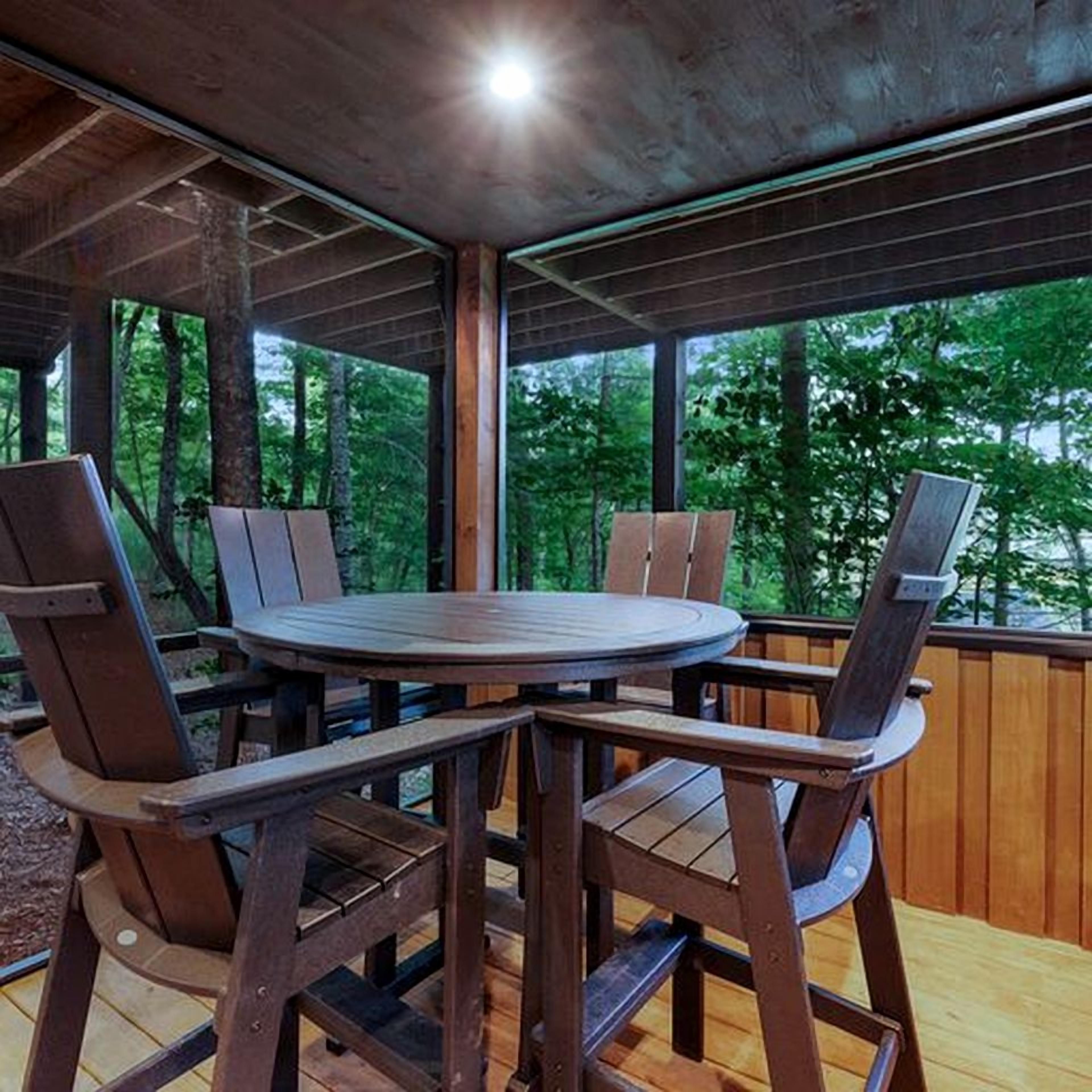 Une terrasse extérieure en bois, avec une table ronde et des chaises, entourée de rampes en bois et d’arbres.