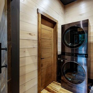 Quarto compacto com porta de madeira, máquina de lavar e secar embutidas e um pequeno banco de madeira.