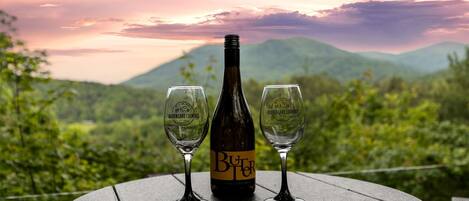 Una botella de vino y dos copas sobre una mesa, con una vista panorámica de montañas y un atardecer.