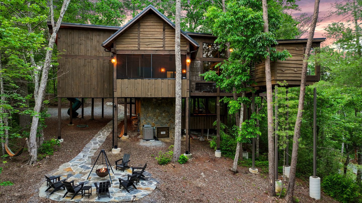 A wooden cabin with a stone fire pit and outdoor seating, surrounded by trees.