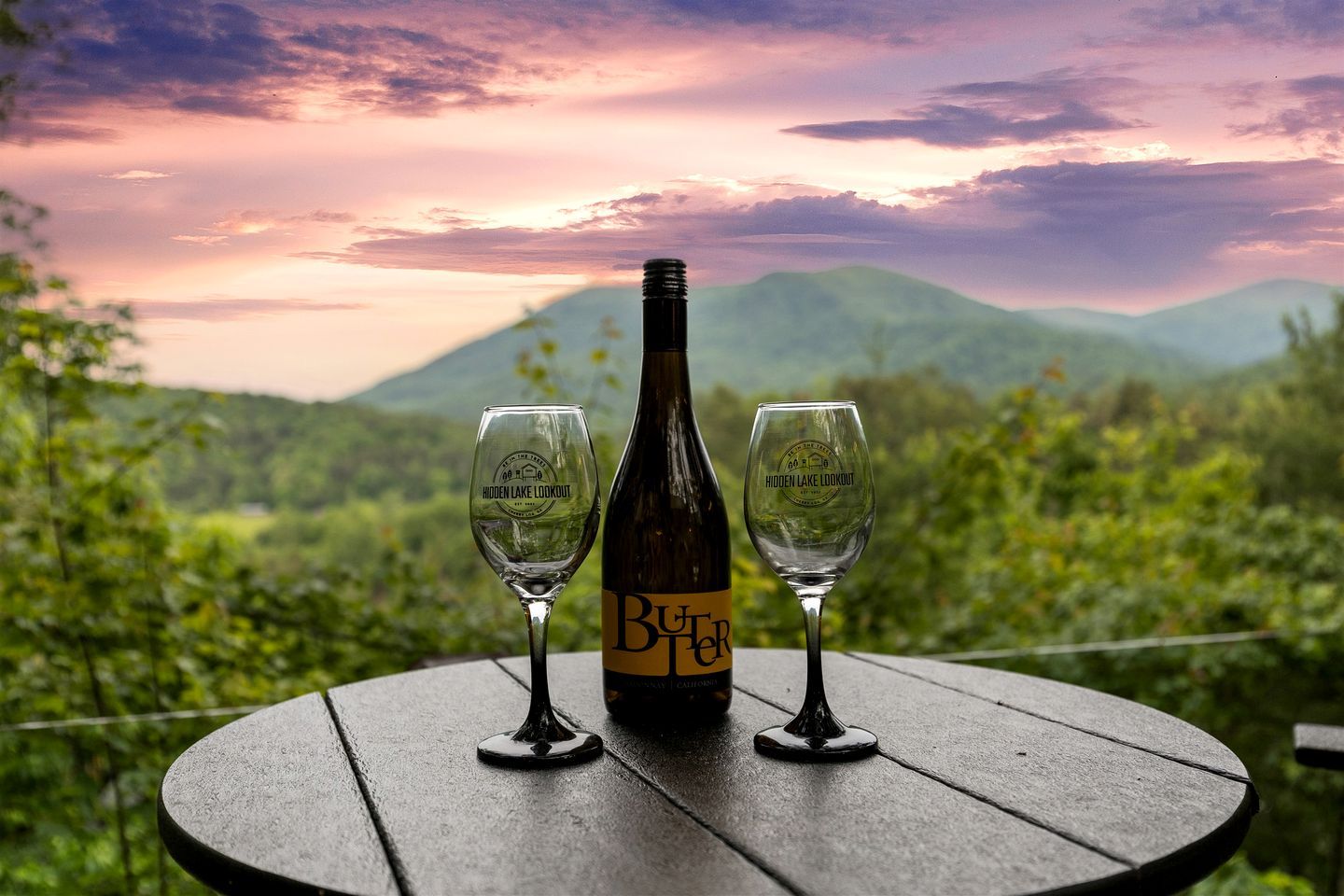 A bottle of wine and two glasses on a table with a scenic view of mountains and a sunset.