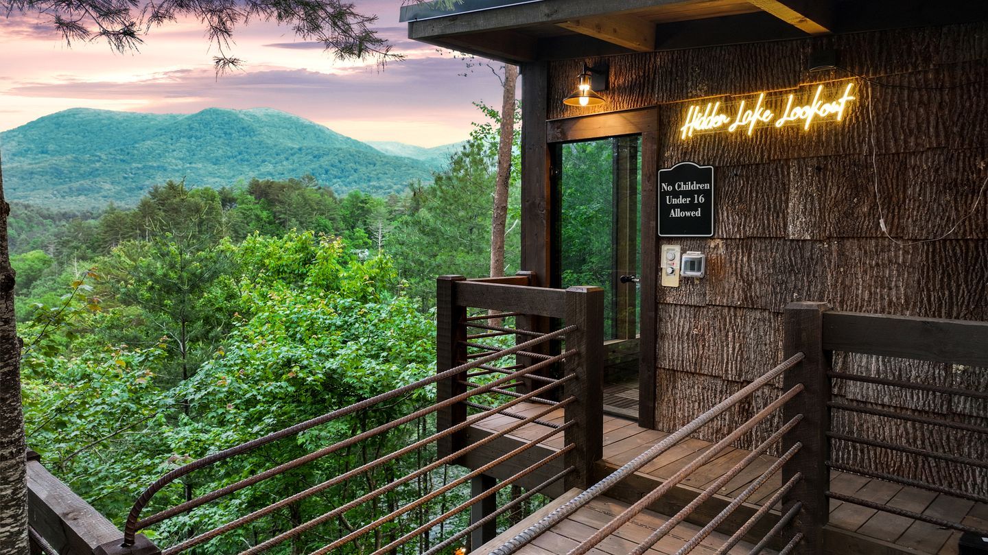 A wooden deck with a view of a forested mountain range, a neon sign reading 'Hidden Lake Lookout,' and a 'No Children Under 16 Allowed' sign.