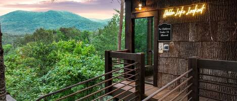 A wooden deck with a view of a forested mountain range, a neon sign reading 'Hidden Lake Lookout,' and a 'No Children Under 16 Allowed' sign.
