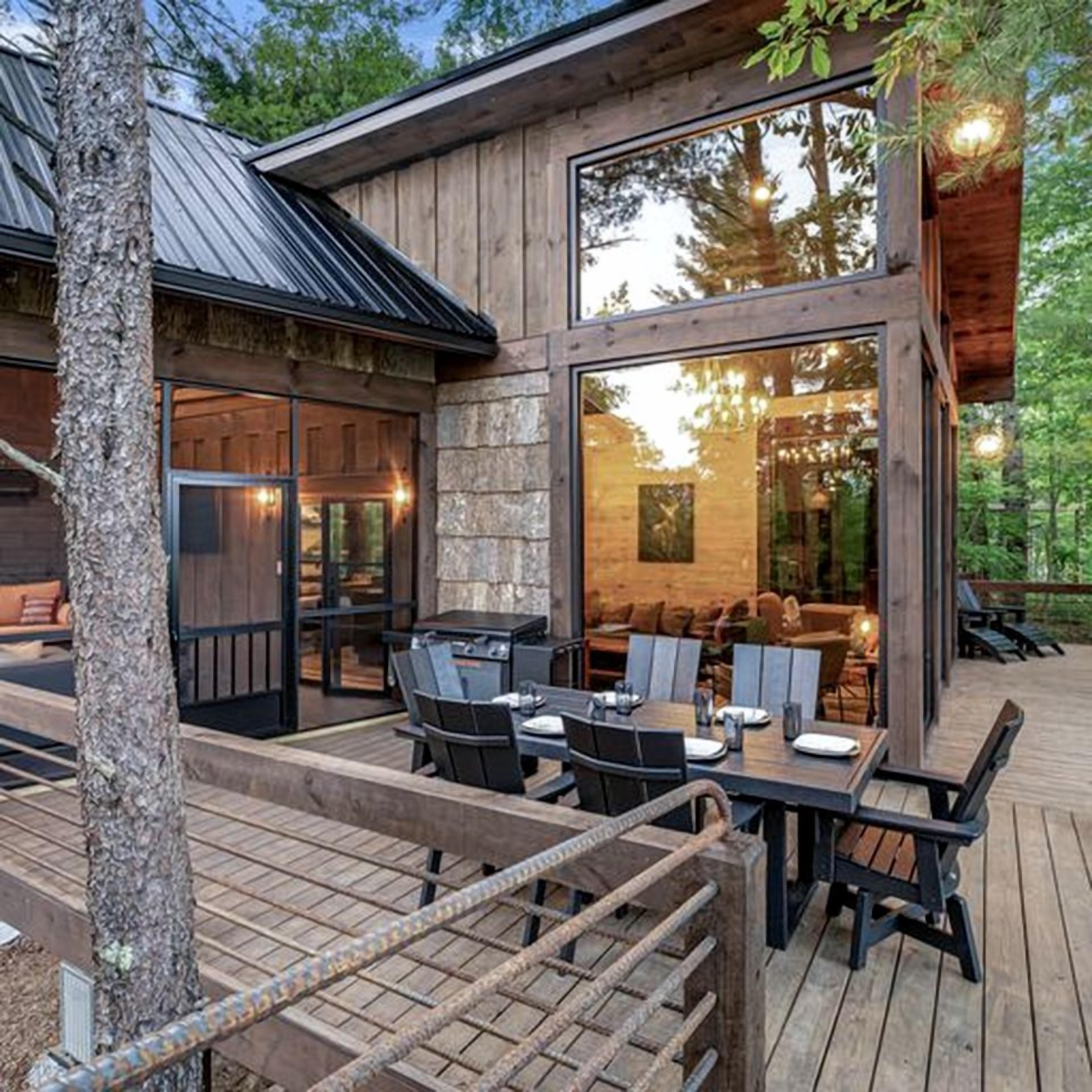 A wooden cabin with a deck, outdoor seating, and a large window overlooking the forest.