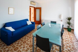 A room with a blue sofa, a glass-top dining table, chairs, a cabinet, a potted plant, and patterned flooring. - Refined Venice Lido Retreat with Terrace, Garden & Steps to the Beach (Lido)