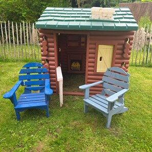 Een klein houten gebouw met een groen dak, een deur en een raam. Twee Adirondack-stoelen staan ervoor.