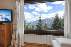 Ein Zimmer mit Fernseher auf einer Holzkommode, einem großen Fenster mit Blick auf die Berge und einem weißen Sofa.