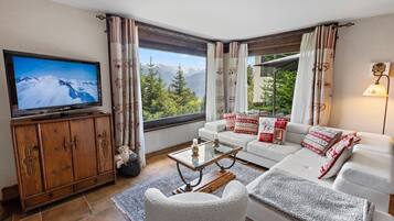 Un salon équipé d’un téléviseur, d’un canapé, d’une table basse et offrant une vue sur les montagnes grâce à de grandes fenêtres.