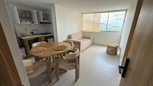 A modern kitchen with built-in appliances, a dining area with a wooden table and chairs, a sofa, and a large window with a city view. - Premium studio apartment on the Golden Mile (ibague)