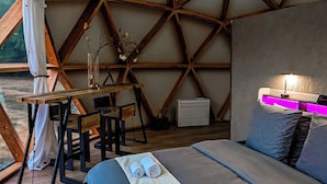 A room with a wooden ceiling structure, a bed with a grey and white bedspread, a desk with a chair, and a table with a lamp.