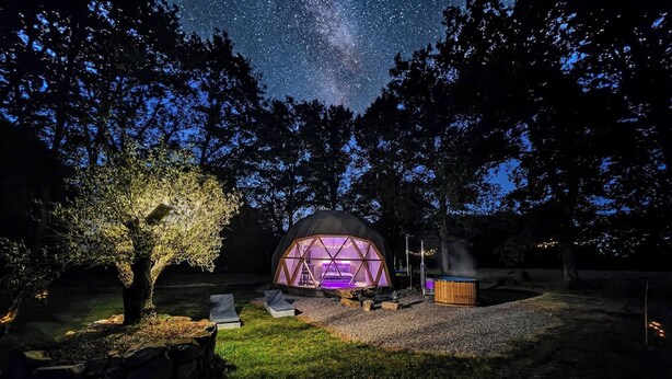 A geodesic dome structure with a central seating area and a hot tub, surrounded by trees and a starry sky.