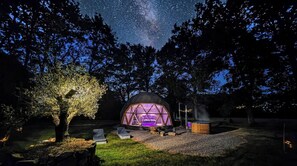 Une structure en forme de dôme géodésique dotée d’un espace central aménagé en salon et d’un jacuzzi, le tout entouré d’arbres et surplombé par un ciel étoilé.