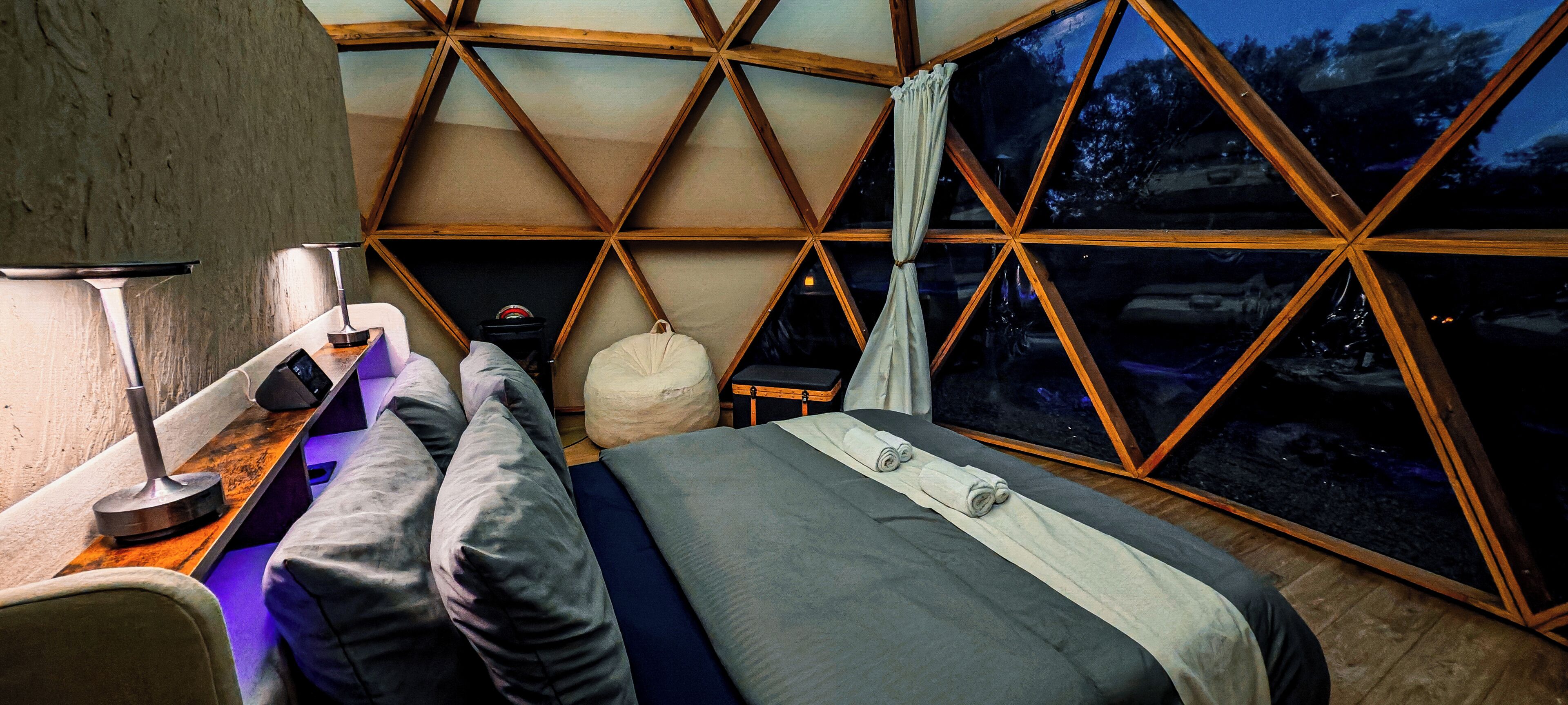Een kamer met een bed, nachtkastje, lamp en raam met uitzicht op de nachthemel.