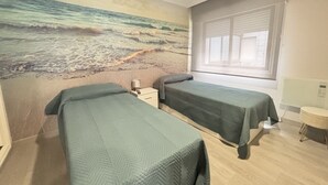 Quarto com duas camas, um mural de praia, uma janela com persianas e uma pequena mesa de cabeceira com abajur.