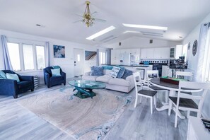 A modern living room with a glass coffee table, a sofa, and a dining area with white chairs. - Welcome to The Coastal Crown, your ultimate luxury beach getaway! 👑<br>✨ Top-Floor Penthouse<br>🏖️ Steps to the Beach<br>🌊 Balcony Ocean Views<br>🛁 Master Suite Jacuzzi<br>📺 85" TV & Skylights (North Myrtle Beach)