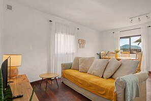 A living room with a sofa, a small round table, and a lamp. There is a window with curtains and a view of the outdoors. - La Casita II in Masdache: Tranquility in the Heart of Lanzarote (Masdache)