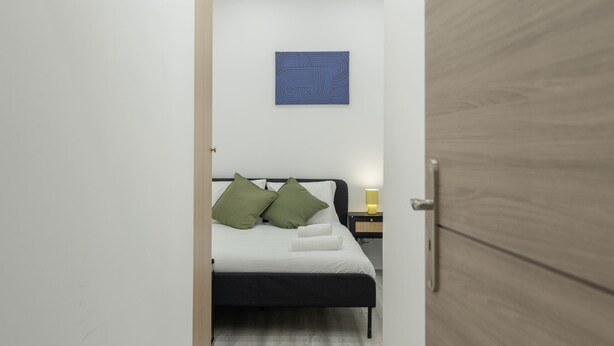 Un dormitorio con cama, almohadas, una mesita de noche con lámpara y una obra de arte en la pared.