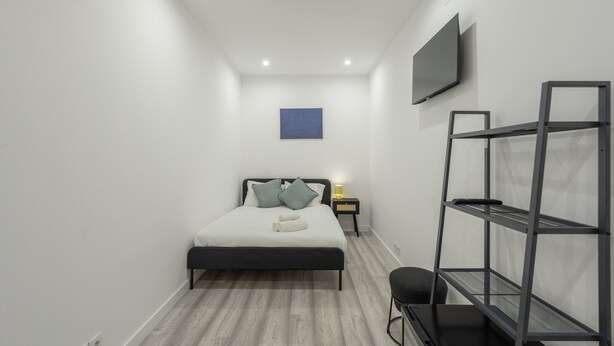 A bedroom with a bed, a TV mounted on the wall, a nightstand with a lamp, and a shelving unit. - Lisbon Modern Suites IV by Homing (Lisbon)