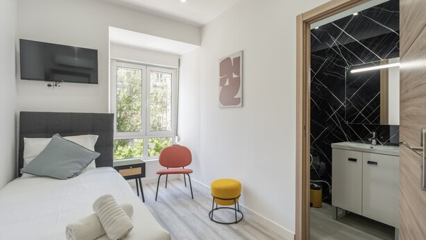 Ein modernes Hotelzimmer mit Bett, Fernseher, Fenster, Stuhl, Hocker und Bad.