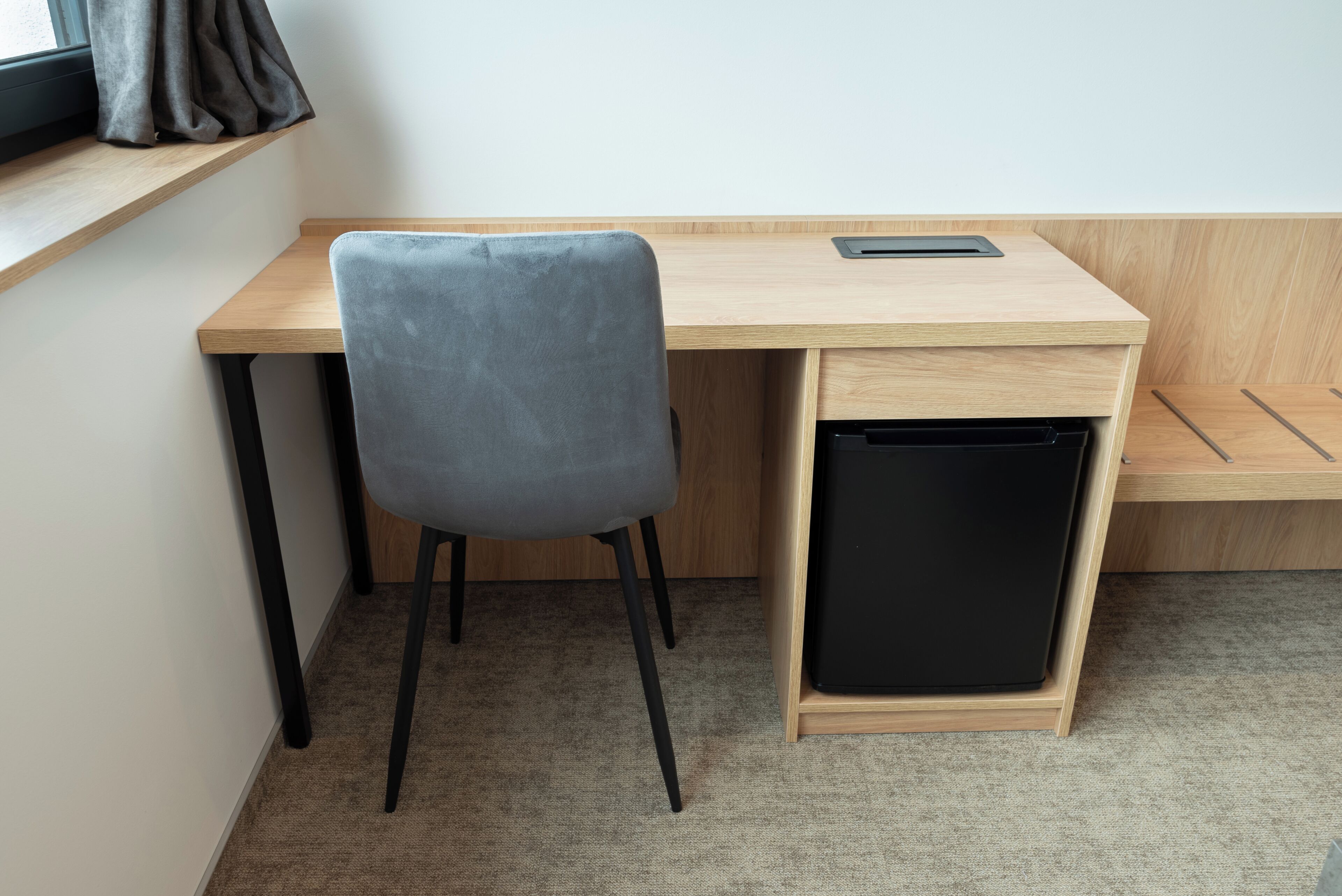 A desk with a chair, a black trash bin, and a window with curtains.