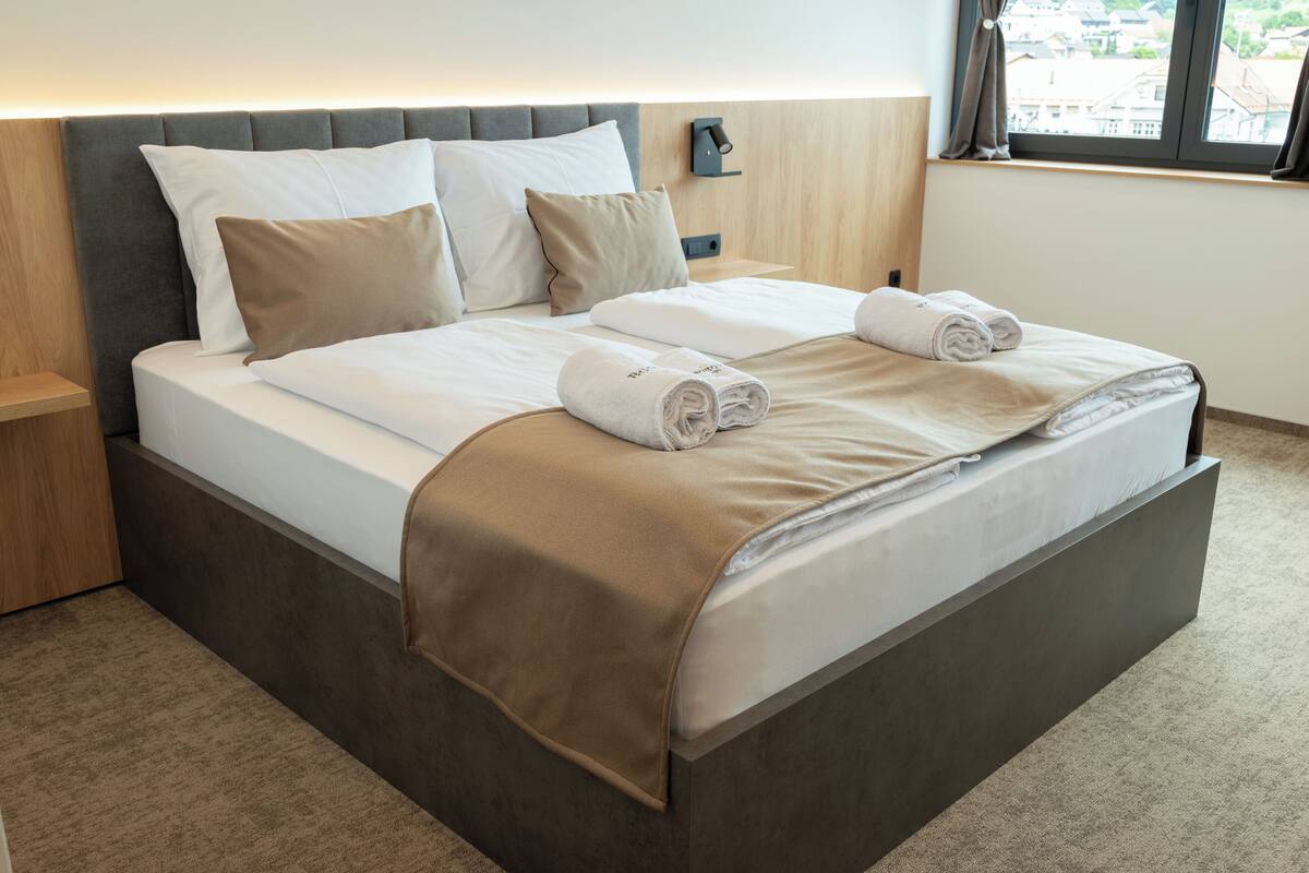 A neatly made bed with a grey headboard, white linens, and two rolled towels.