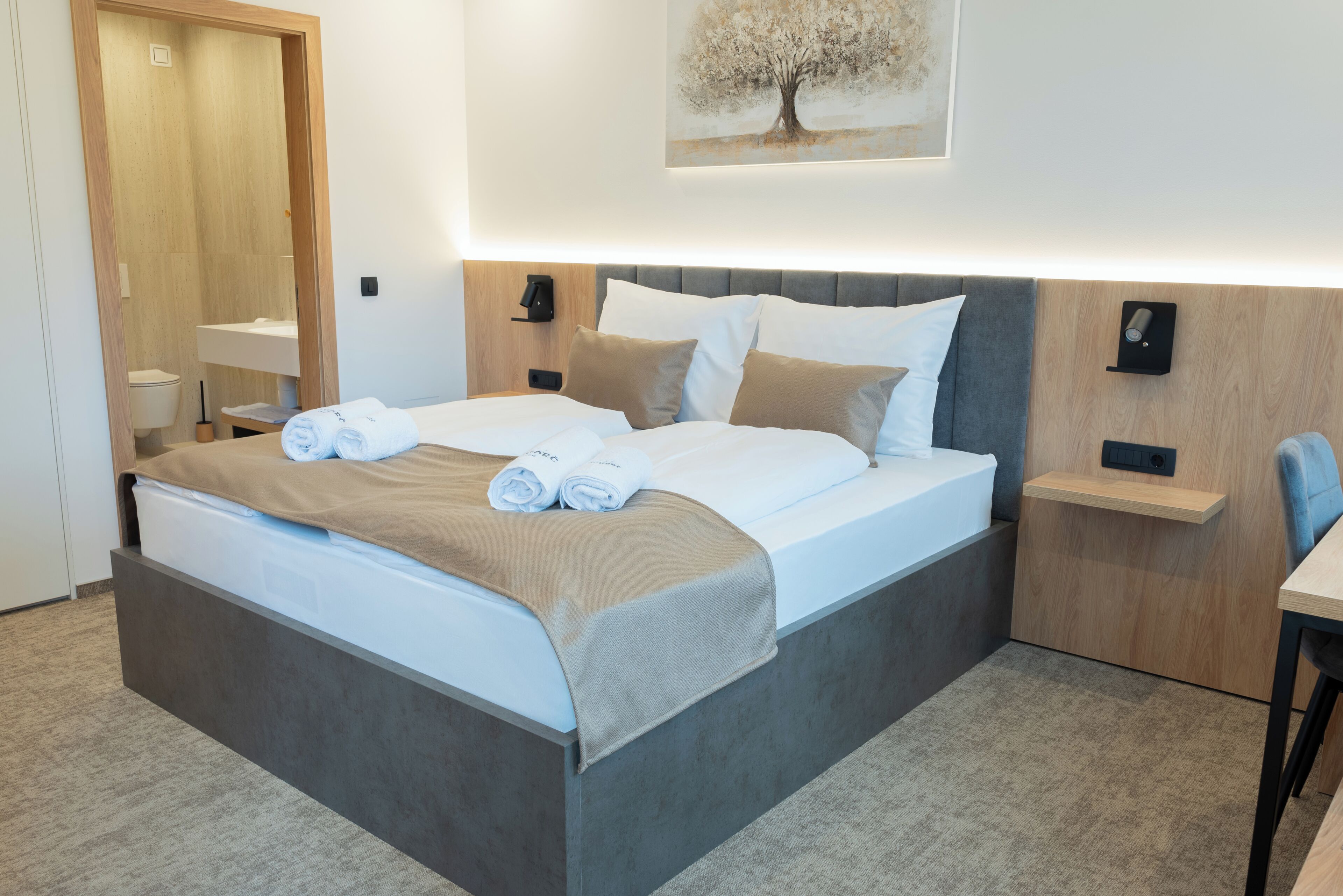 Una habitación de hotel moderna con una cama grande, una mesita de noche, una lámpara de pared y un cuadro de un árbol.