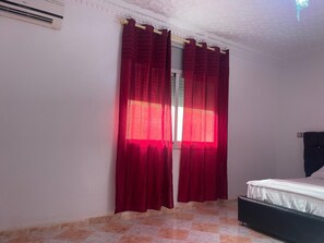 Un dormitori amb un llit, una finestra amb cortines vermelles i un aparell d'aire condicionat a la paret.