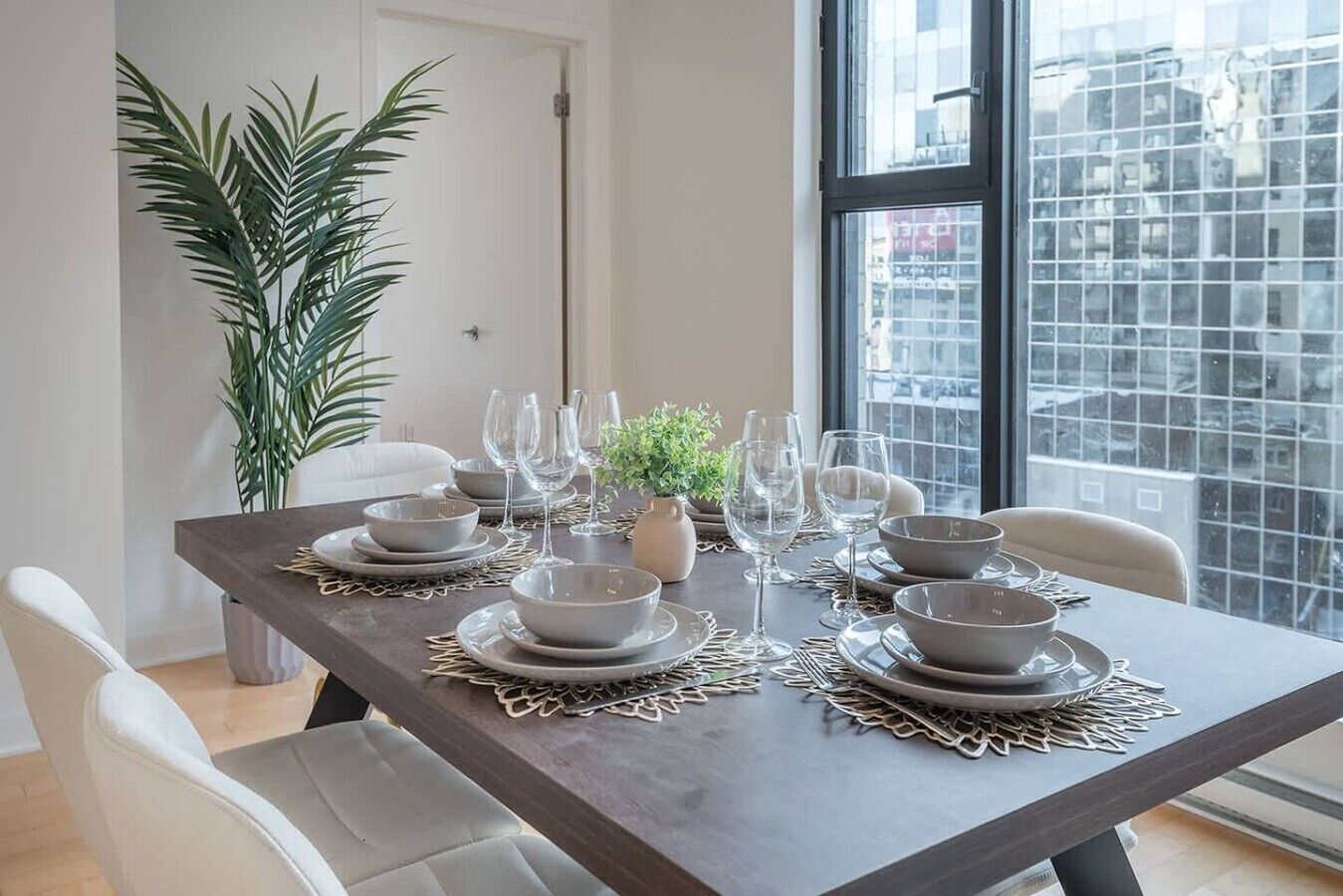 Een eettafel gedekt met borden, kommen en wijnglazen, omringd door stoelen, met een groot raam en een kamerplant op de achtergrond.
