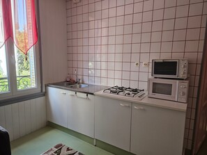 Une cuisine avec des meubles blancs, une cuisinière à gaz, un four à micro-ondes et une fenêtre ornée d’un rideau rouge.