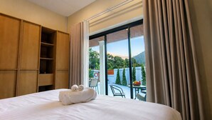 Habitación de hotel con cama, armario de madera, balcón con mesa y sillas, y vista a zonas verdes y montañas.
