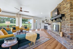 A spacious living room with a stone fireplace, yellow sofas, and a large window overlooking a lawn. - The Back Nine is your Hot Springs golf course getaway! (Hot Springs)