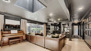 A spacious living area with a piano, a large window, a sofa, and a kitchen in the background. - Urban House in the Trees (Dallas)