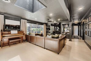 A spacious living area with a piano, a large window, a sofa, and a kitchen in the background. - Urban House in the Trees (Dallas)