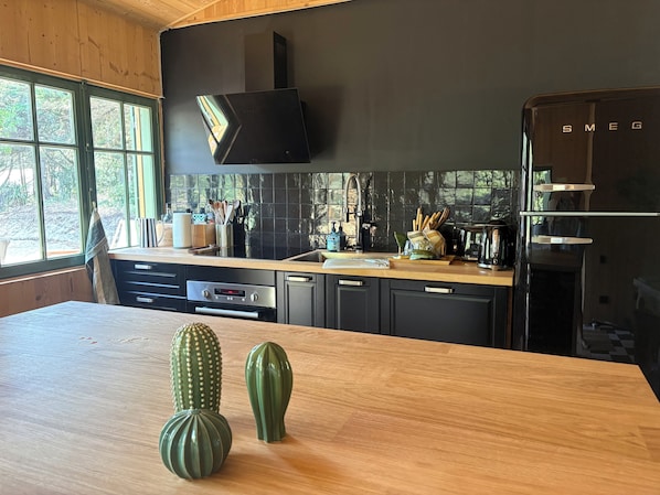 A modern kitchen with a wooden countertop, black cabinets, a black refrigerator, and a window with a view of trees. - House at the Foot of the Dune (L ège-Cap-Ferret)