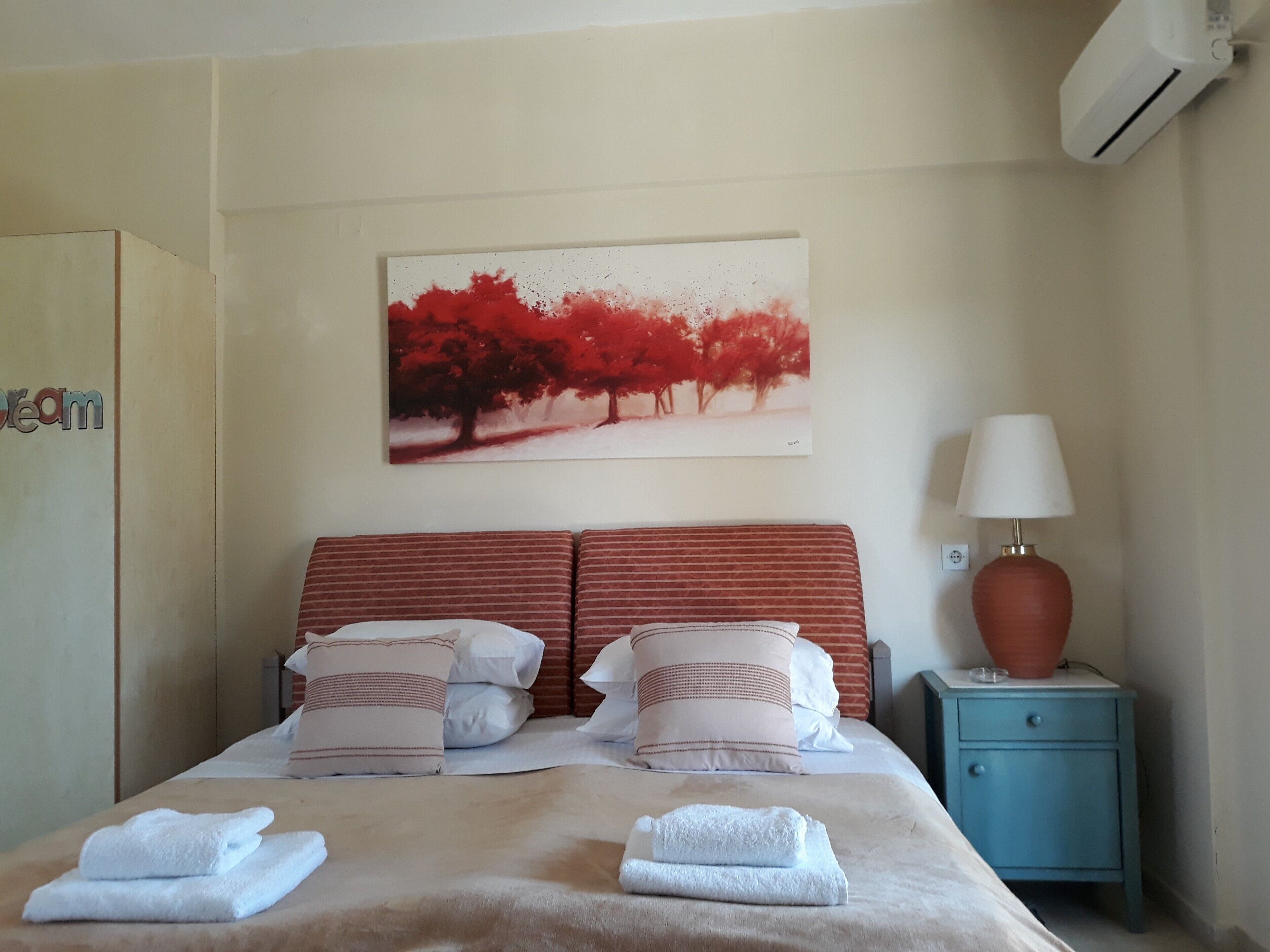 A hotel room with a bed, pillows, a nightstand with a lamp, a wall-mounted air conditioner, and a painting of trees.