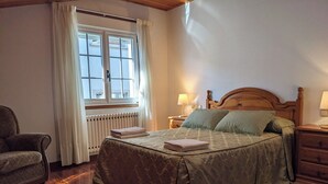 Ein Schlafzimmer mit einem hölzernen Bett, einem Sessel, einem Nachttisch mit Lampe, einem Fenster mit Vorhängen und einer Heizung.