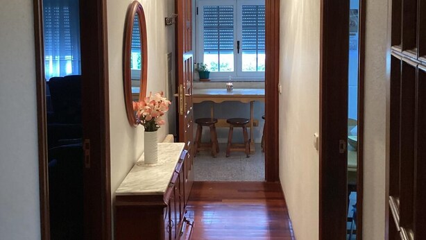 Ein schmaler Korridor mit Holzboden, einem Spiegel links, einem kleinen Tisch mit Blumenvase und einem Fenster mit Jalousien im Hintergrund.