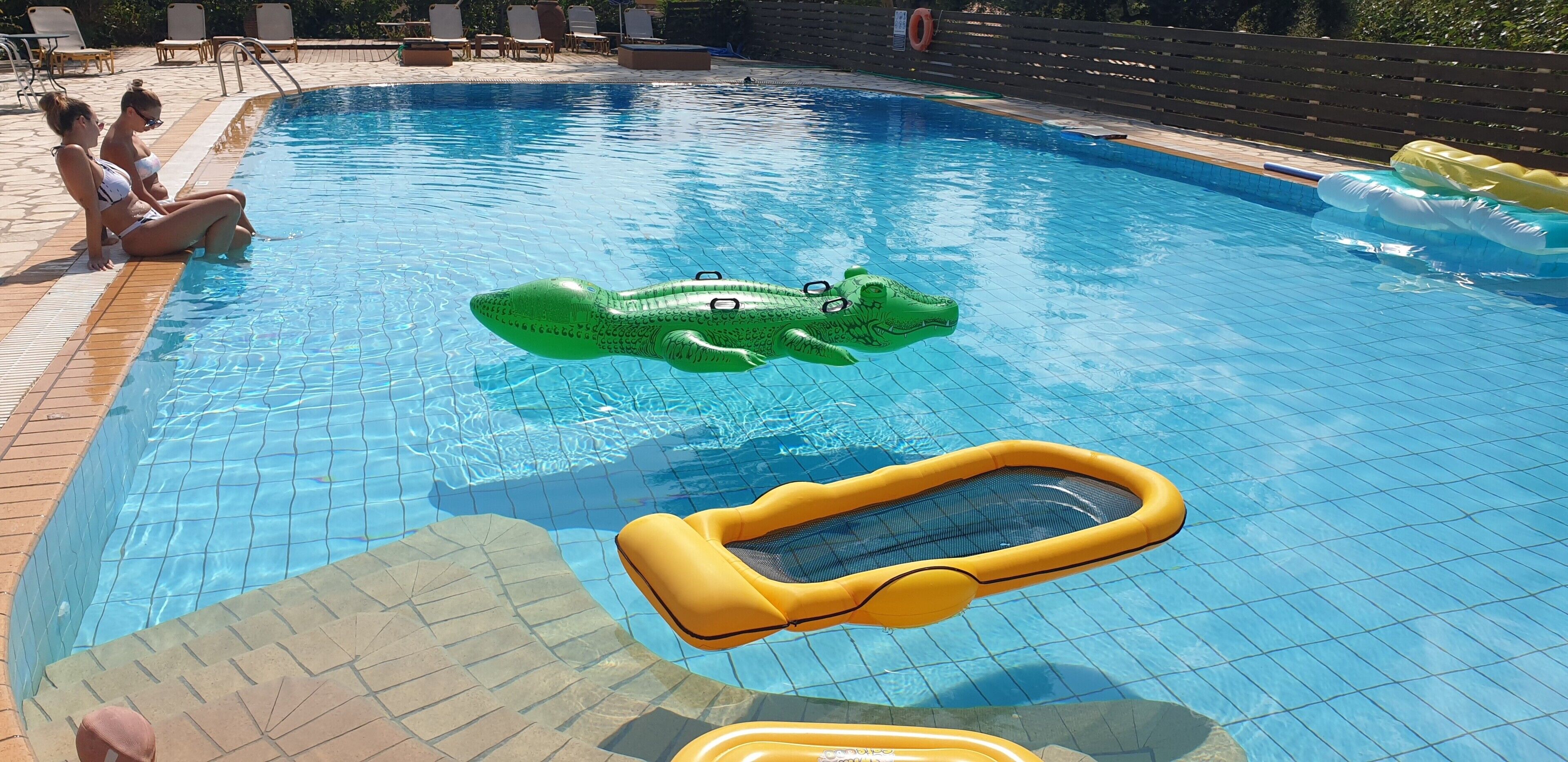 Uma piscina com duas pessoas sentadas na borda, um flutuador de crocodilo inflável verde e um flutuador de piscina amarelo.