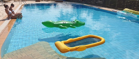 Uma piscina com duas pessoas sentadas na borda, um flutuador de crocodilo inflável verde e um flutuador de piscina amarelo.