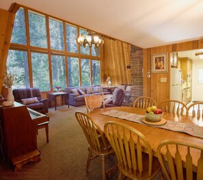 Ein gemütliches Wohnzimmer mit einem Klavier, einem Esstisch mit Stühlen, einer Couch und einem großen Fenster mit Blick auf Bäume.