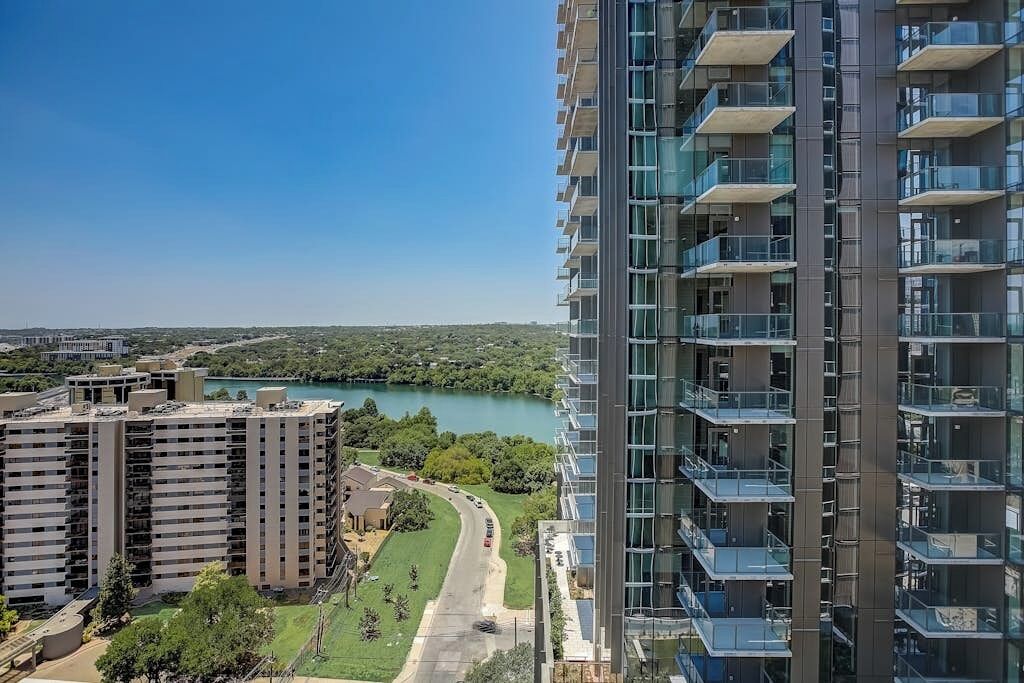 A high-rise building with multiple balconies, a river in the background, and a road leading to a parking area.
