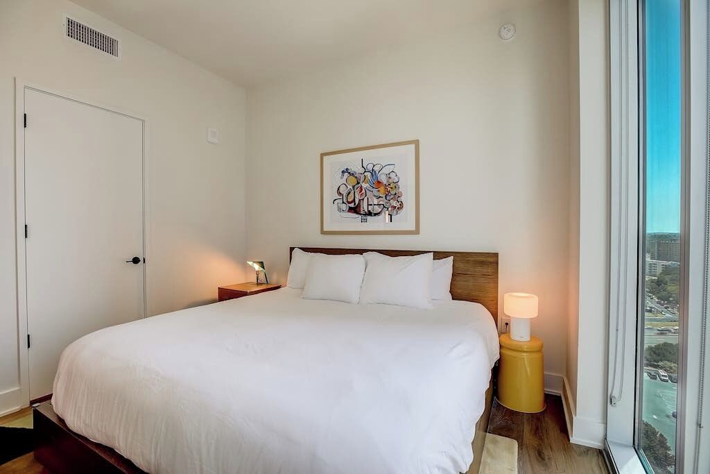 A hotel room with a large bed, a framed abstract painting, a bedside lamp, and a window with a city view.
