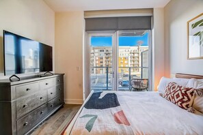 Et soveværelse med seng, kommode, fjernsyn og balkon med udsigt til bygninger.