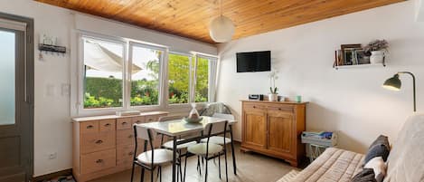 A compact living space with a dining area, a sofa, a TV, and a view of greenery through the windows.