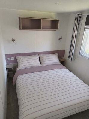 Ein Bett mit Kopfteil, ein Nachttisch, ein Fenster mit Vorhängen und ein an der Wand befestigtes Regal.