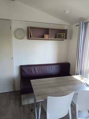 Ein Zimmer mit einem Holztisch, weißen Stühlen, einem dunkelgrauen Sofa, einer Wanduhr und einem Regal mit unterschiedlichen Gegenständen.