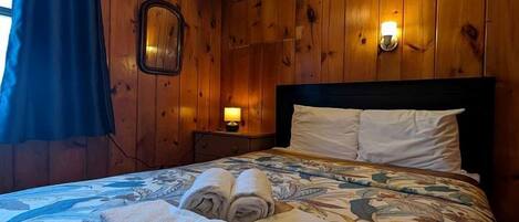 A wooden-paneled room with a bed, a nightstand, and a wall-mounted light fixture.